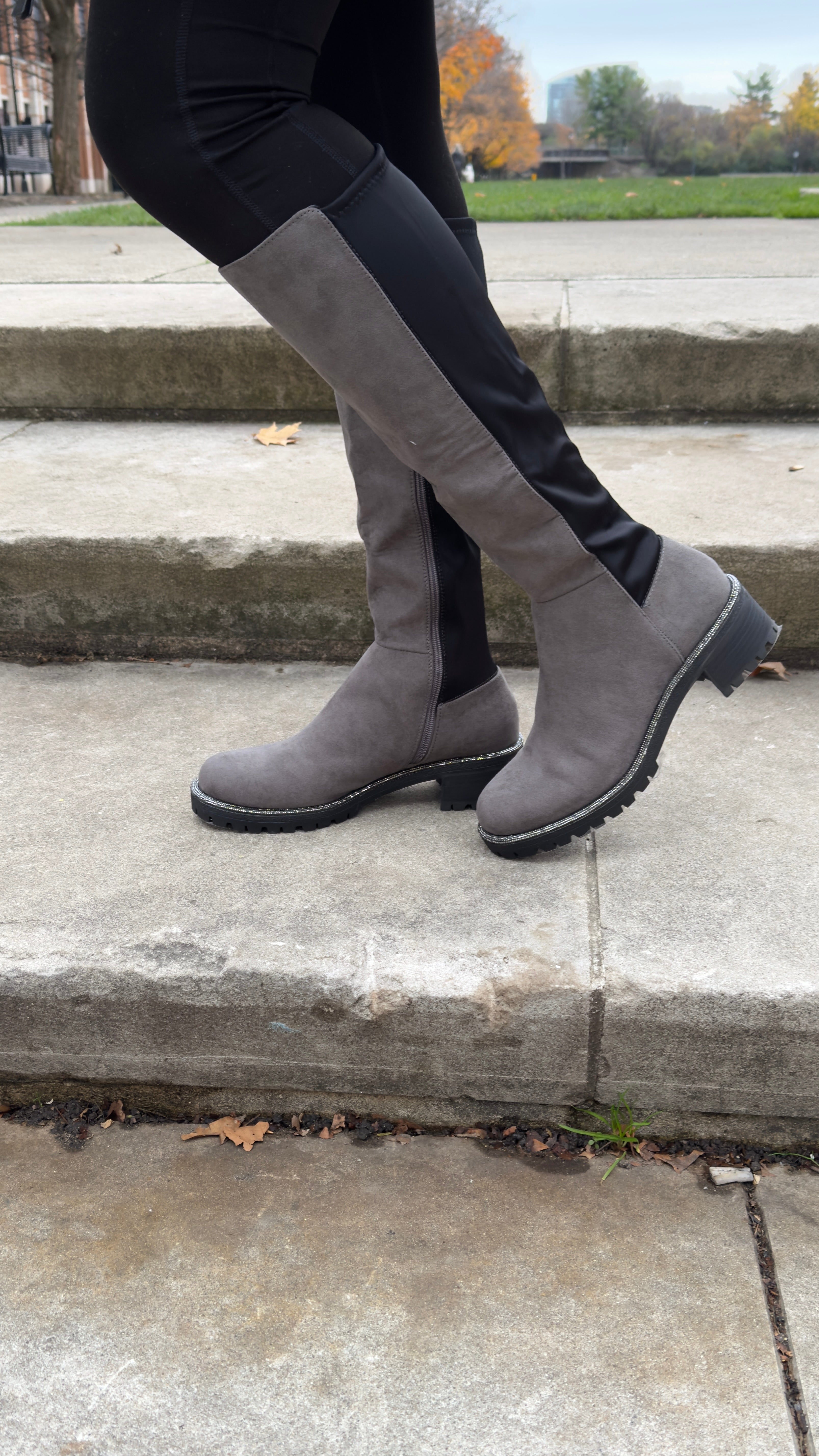 A woman stands on outdoor concrete steps wearing gray knee-high suede boots with black stretch panels and low lug soles. The boots pair stylishly with black leggings, creating a sleek and comfortable fall look perfect for everyday wear.